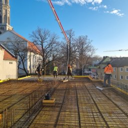 AJON SPÓŁKA Z OGRANICZONĄ ODPOWIEDZIALNOŚCIĄ - Zbrojenie stropu budynku w trakcie betonowania. Widoczni pracownicy w odblaskowych kamizelkach i pompa do betonu. W tle zabudowa miejska i wieża kościoła.