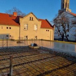 AJON SPÓŁKA Z OGRANICZONĄ ODPOWIEDZIALNOŚCIĄ - Zalewanie betonem stropu budynku z widoczną siatką zbrojeniową i wystającymi prętami. W tle budynek z czerwoną dachówką i wieża kościoła w porannym słońcu.