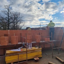 AJON SPÓŁKA Z OGRANICZONĄ ODPOWIEDZIALNOŚCIĄ - Murarz w pracy: wznoszenie ściany z czerwonej cegły na rusztowaniu. Widoczne narzędzia, materiały budowlane i fragment krajobrazu miejskiego w tle.