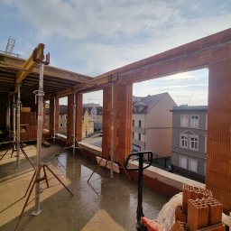AJON SPÓŁKA Z OGRANICZONĄ ODPOWIEDZIALNOŚCIĄ - Wznoszenie ścian z czerwonej cegły ceramicznej na budowie. Widoczne podpory stropowe, paleta z cegłami i wózek paletowy. W tle zabudowa miejska.