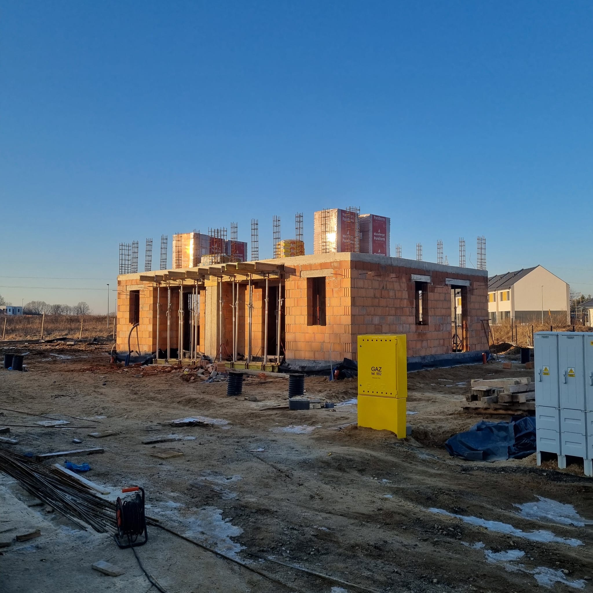 Budowa domu jednorodzinnego w stanie surowym otwartym, z widocznymi elementami zbrojenia i pustakami Porotherm. Na pierwszym planie żółta skrzynka gazowa.
