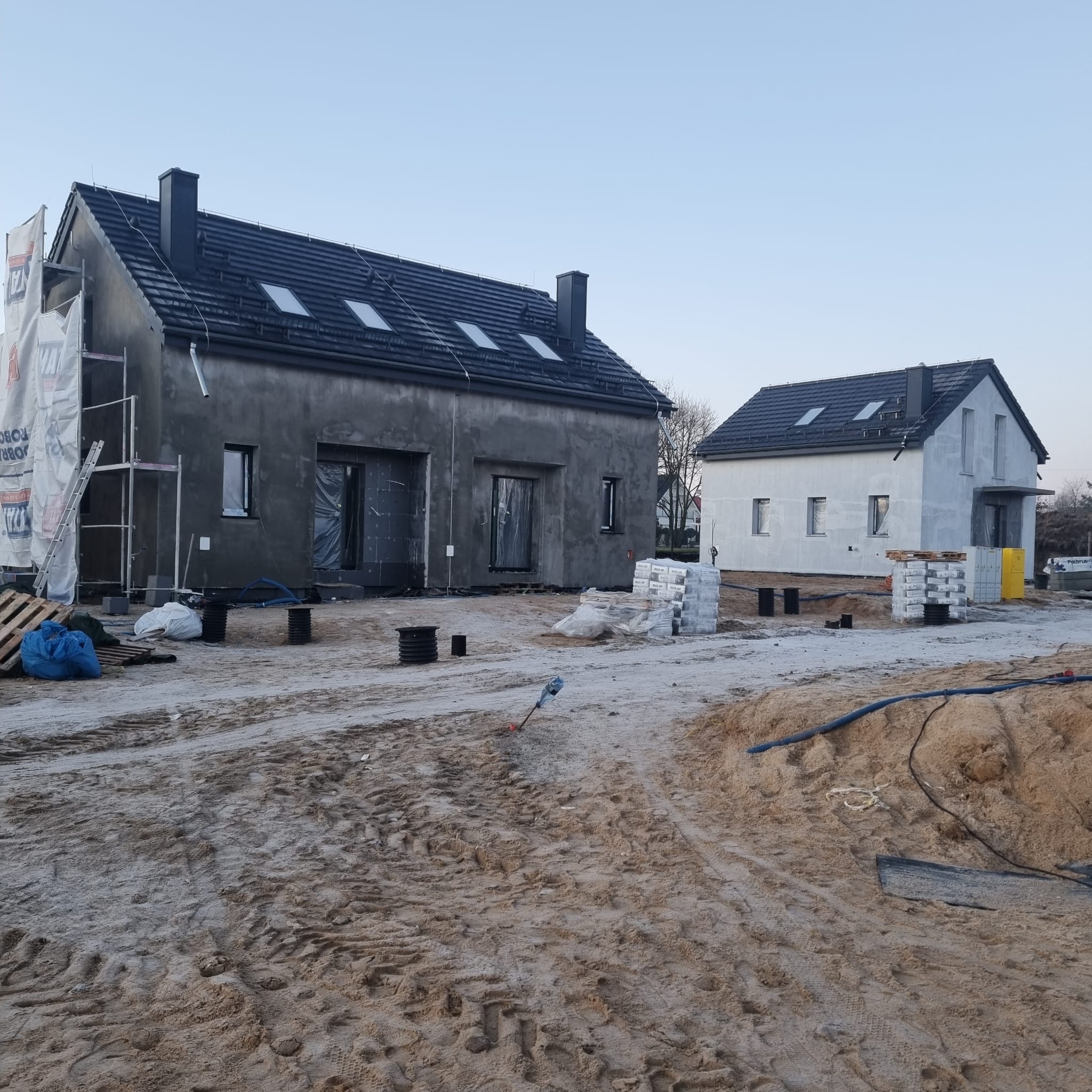 Dwa domy w różnej fazie budowy: jeden z szarą elewacją w trakcie prac, drugi z białą, prawdopodobnie po malowaniu. Na pierwszym widoczne rusztowania i materiały budowlane.