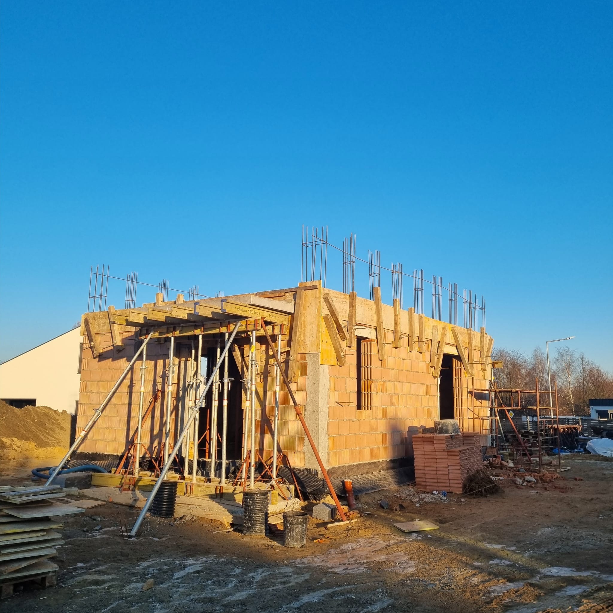 Wznoszenie ścian z pustaków ceramicznych na budowie domu jednorodzinnego. Widoczne szalunki i zbrojenie stropu. Błękitne niebo w tle.