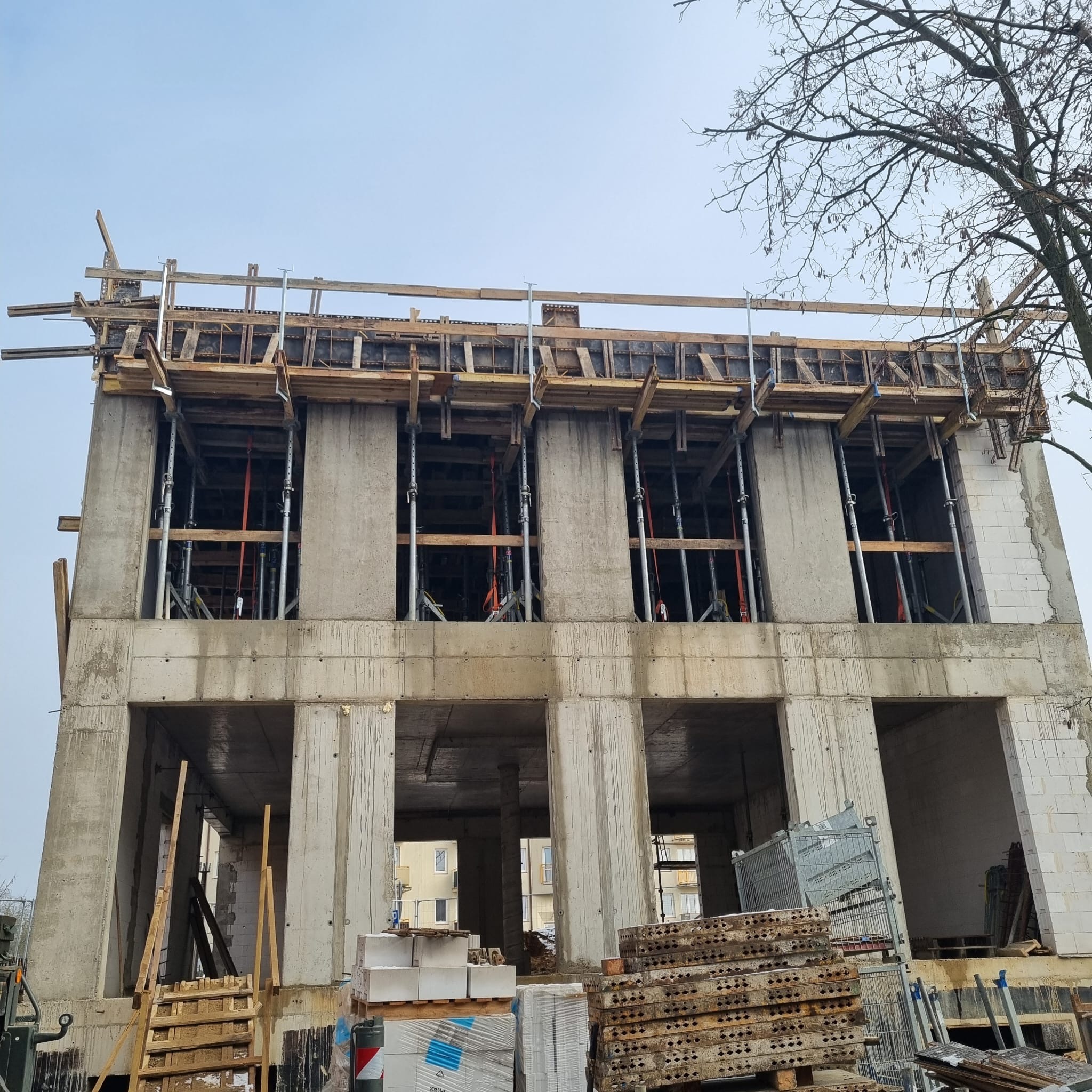 Dwukondygnacyjny budynek w stanie surowym otwartym z widocznym szalunkiem na stropie, materiałami budowlanymi i drzewem w tle.