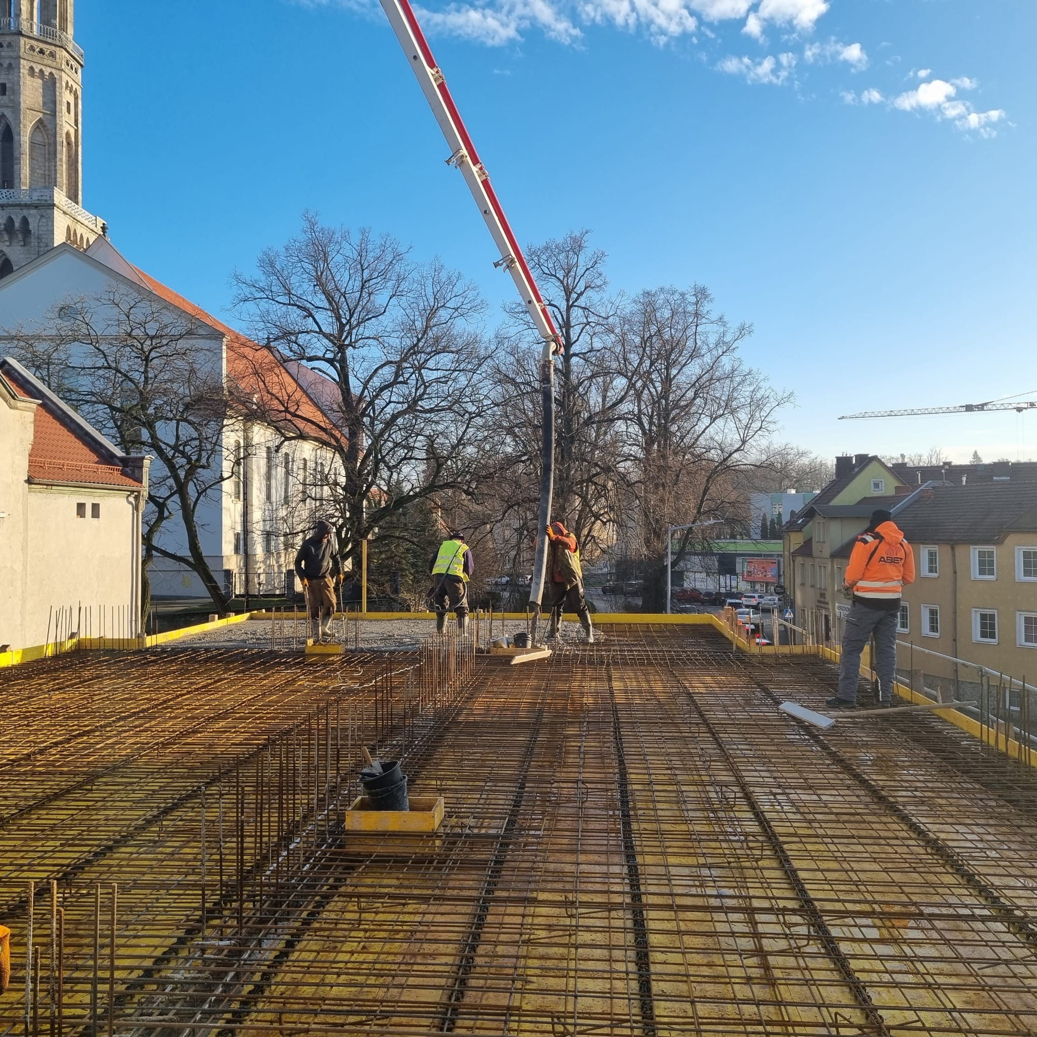 Zbrojenie stropu budynku w trakcie betonowania. Widoczni pracownicy w odblaskowych kamizelkach i pompa do betonu. W tle zabudowa miejska i wieża kościoła.