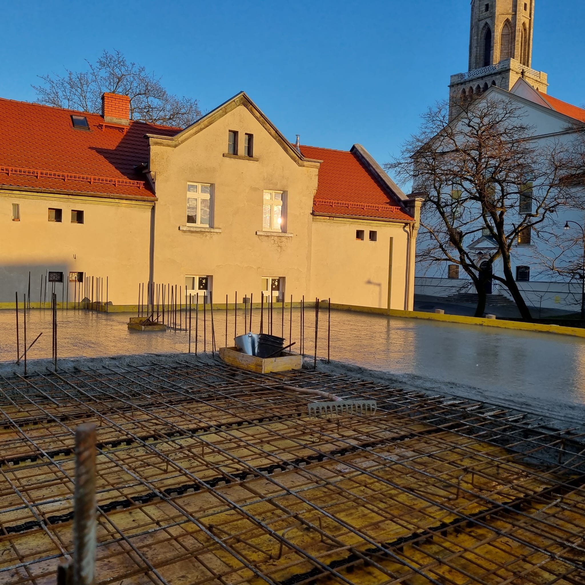 Zalewanie betonem stropu budynku z widoczną siatką zbrojeniową i wystającymi prętami. W tle budynek z czerwoną dachówką i wieża kościoła w porannym słońcu.