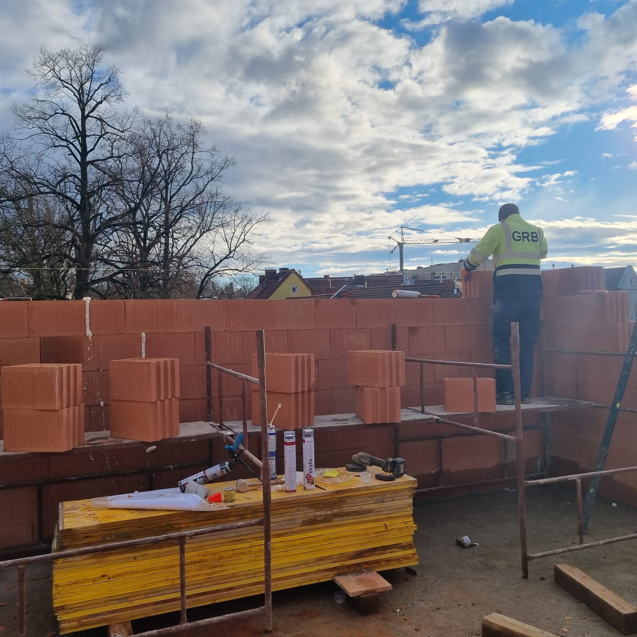 Murarz w pracy: wznoszenie ściany z czerwonej cegły na rusztowaniu. Widoczne narzędzia, materiały budowlane i fragment krajobrazu miejskiego w tle.