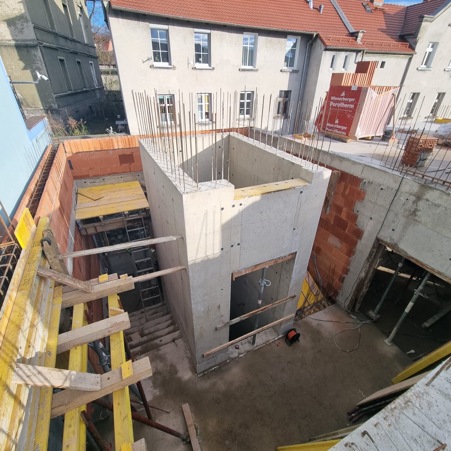 Wysoki, betonowy szyb z otworami, w trakcie budowy. Widoczne zbrojenia, szalunki i cegły Porotherm. Ujęcie z góry, w tle budynki mieszkalne.