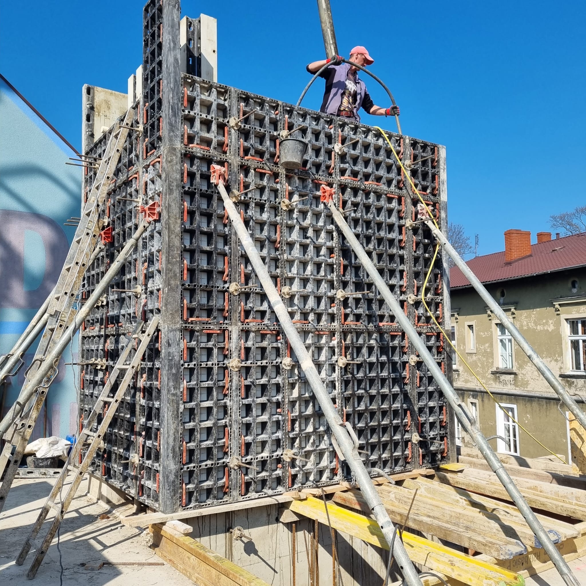 Wznoszenie szalunku z betonu. Robotnik w czerwonej czapce obsługuje pompę do betonu. Konstrukcja wsparta na metalowych podporach. W tle budynki.