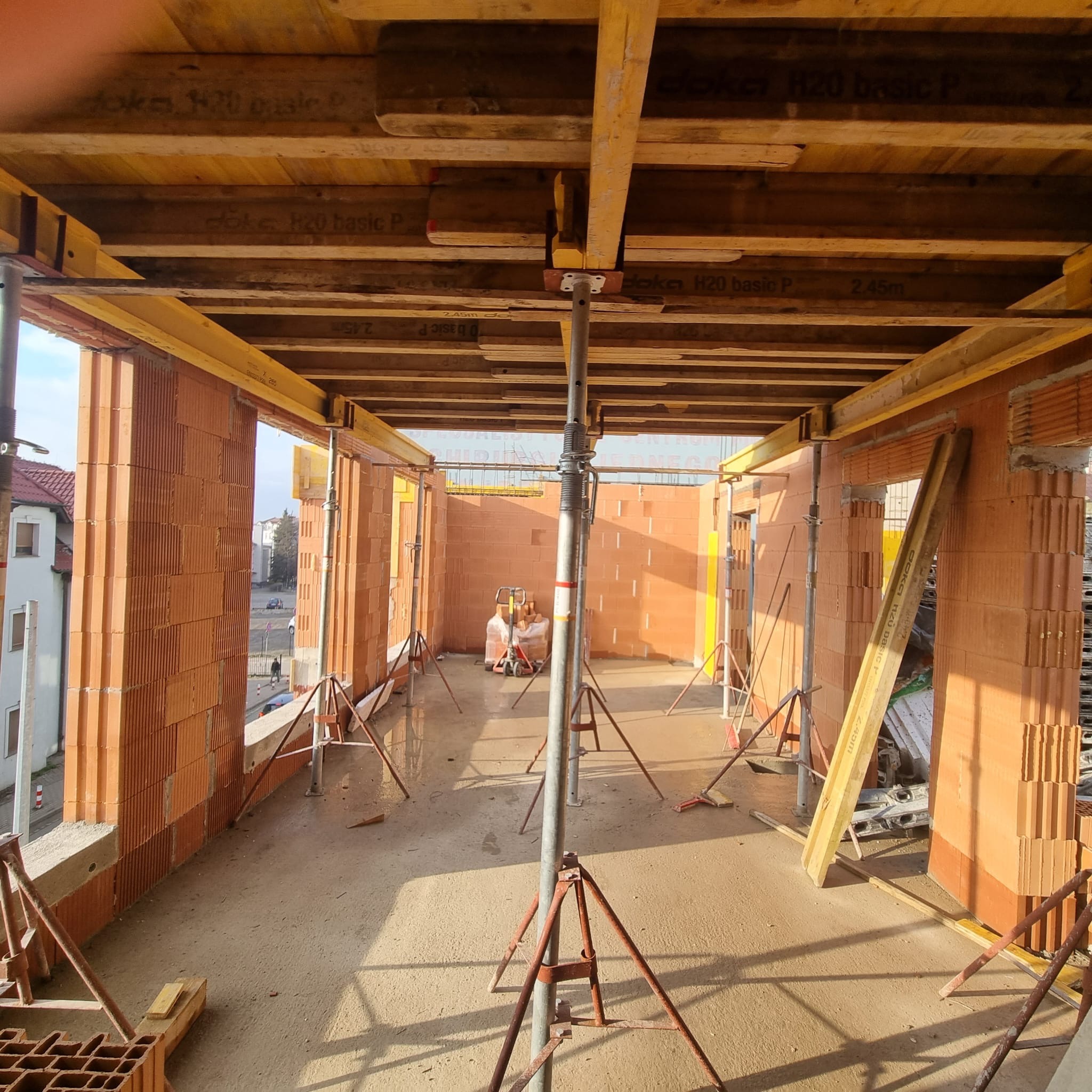 Wnętrze budynku w trakcie budowy. Widoczne ściany z cegły, drewniane szalunki stropu podparte metalowymi stemplami. Na podłodze leżą narzędzia i materiały budowlane.