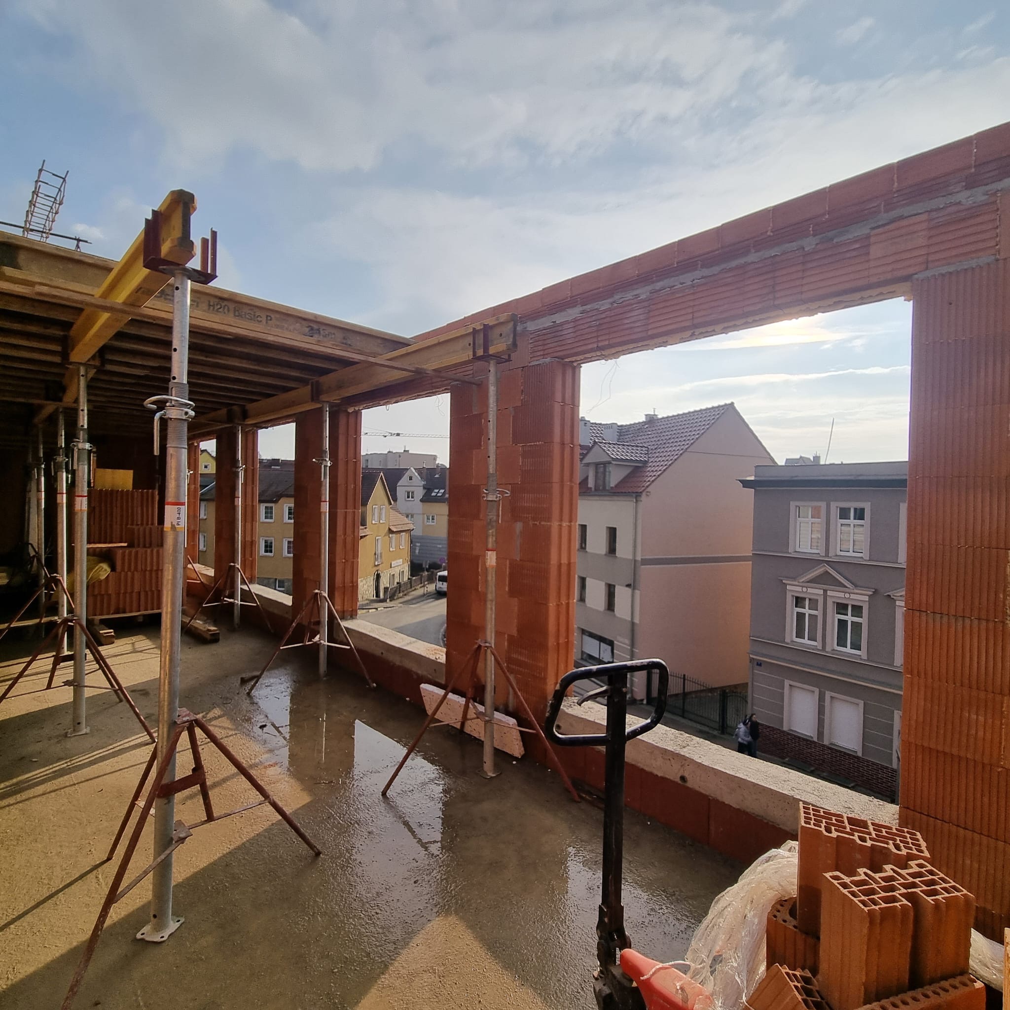 Wznoszenie ścian z czerwonej cegły ceramicznej na budowie. Widoczne podpory stropowe, paleta z cegłami i wózek paletowy. W tle zabudowa miejska.