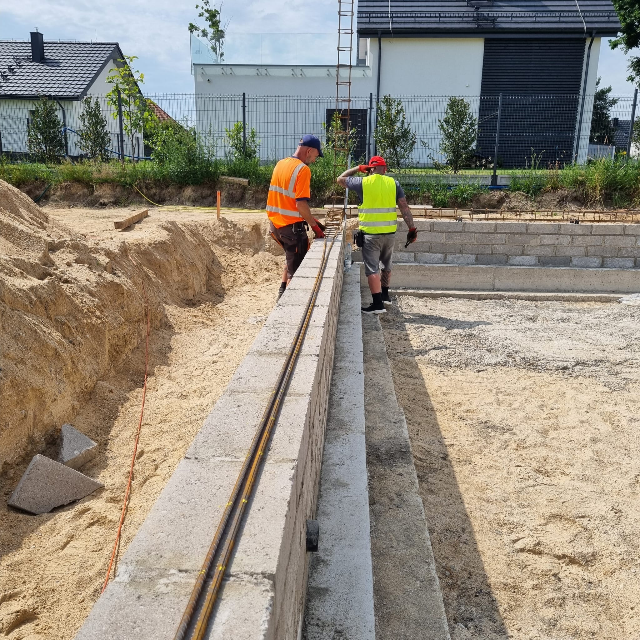 Dwóch pracowników w odblaskowych ubraniach montuje zbrojenie w fundamencie z bloczków betonowych na budowie domu jednorodzinnego w słoneczny dzień.