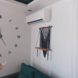 Klima4you Rafał Rabczak - Nowoczesny biały klimatyzator na ścianie w pokoju z zieloną sofą i dekoracyjną półką. Widoczny pilot w dłoni i duży zegar na ścianie.