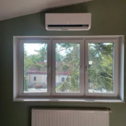 Biała klimatyzacja ścienna nad oknem z widokiem na zieleń, pod oknem biały grzejnik. Ściany w odcieniu zieleni, ukośny sufit.