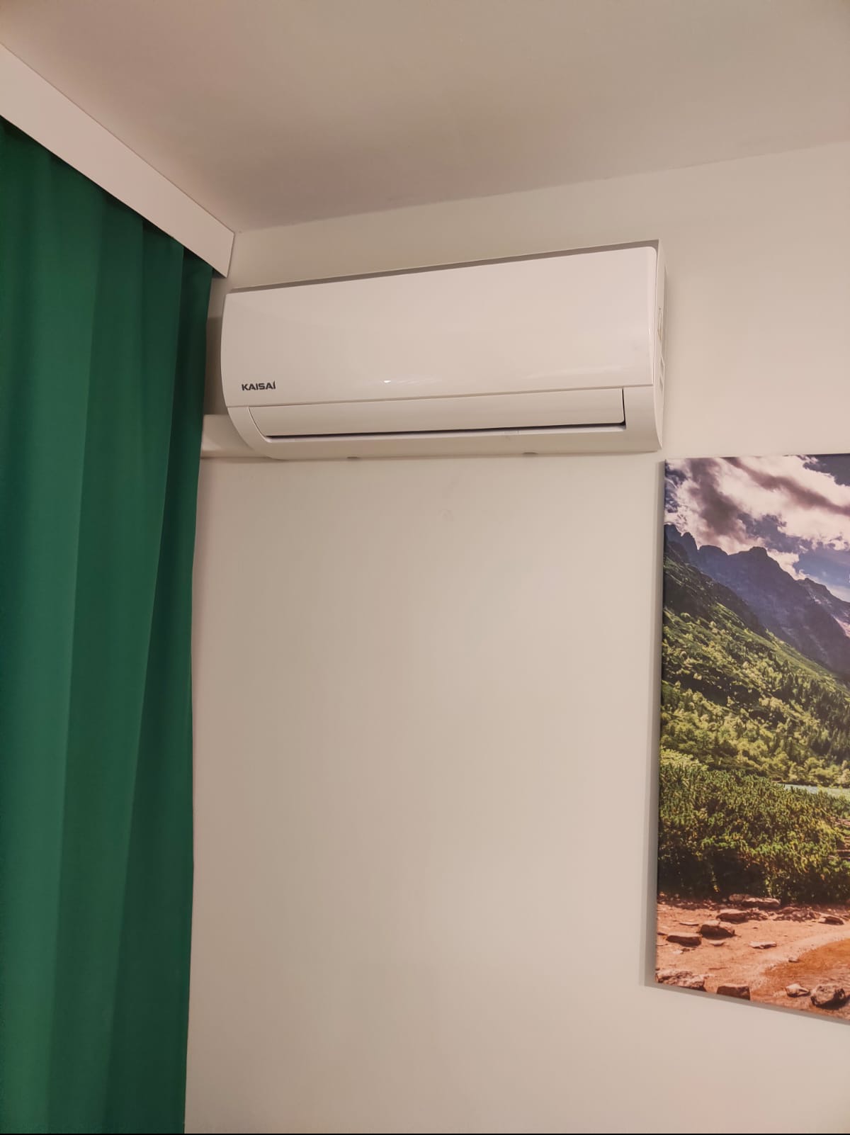 Biały klimatyzator Kaisai na ścianie w pokoju, obok zielona zasłona i obraz z górskim krajobrazem. Widok z dołu, oświetlenie dzienne.