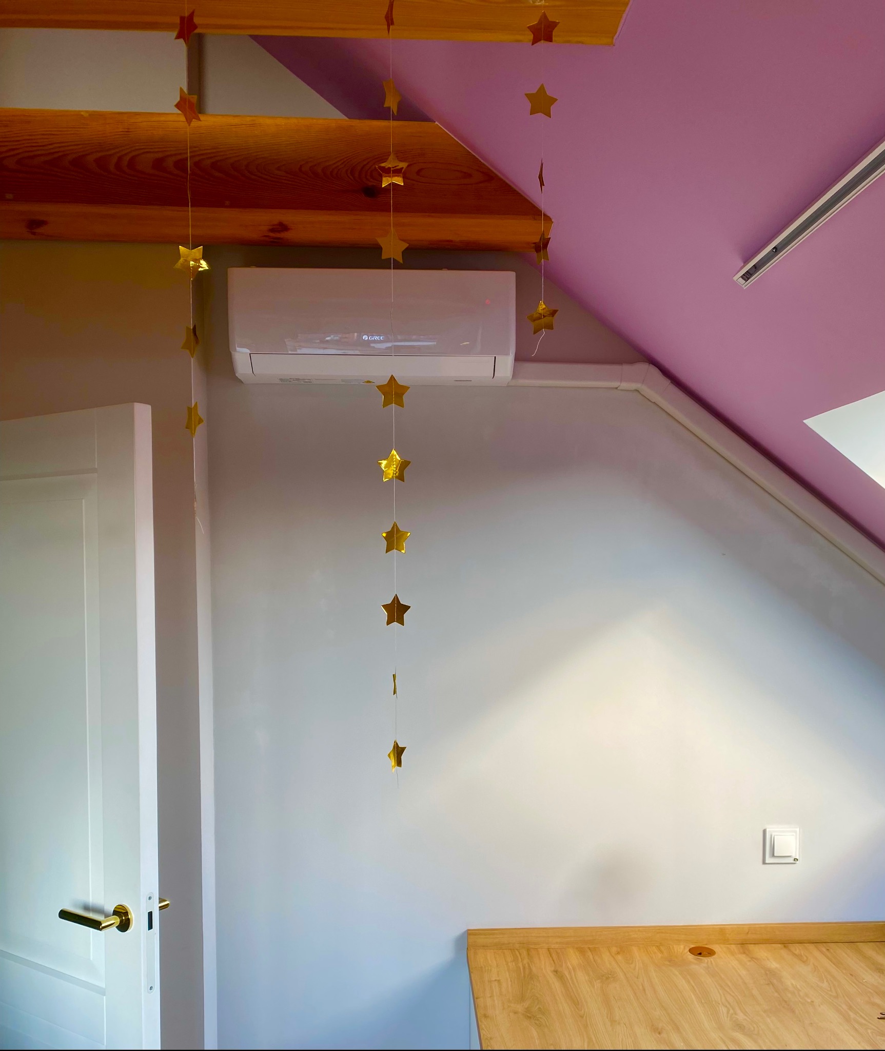 Biały klimatyzator marki Gree zamontowany na szarej ścianie w pokoju na poddaszu, udekorowany złotymi gwiazdkami. Widoczna drewniana belka i fragment biurka.
