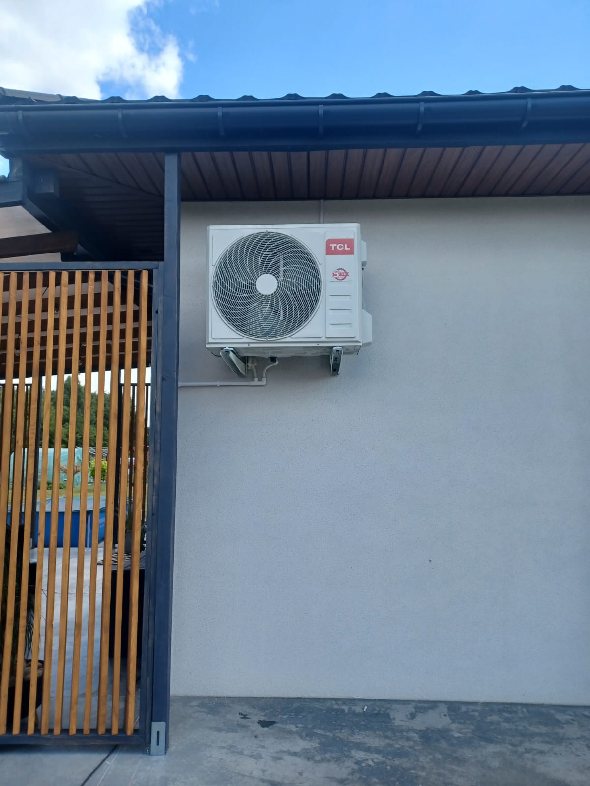 Biała klimatyzacja TCL zamontowana na szarej ścianie budynku. Widoczny fragment drewnianej konstrukcji i niebieskie niebo w tle.