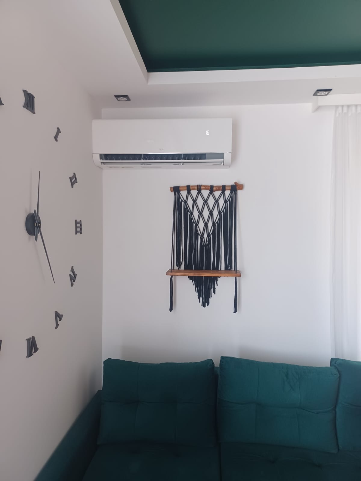 Biały klimatyzator na ścianie w pokoju z dekoracjami: zegar, makrama z półką, zielona sofa i sufit. Nowoczesne wnętrze z klimatyzacją.