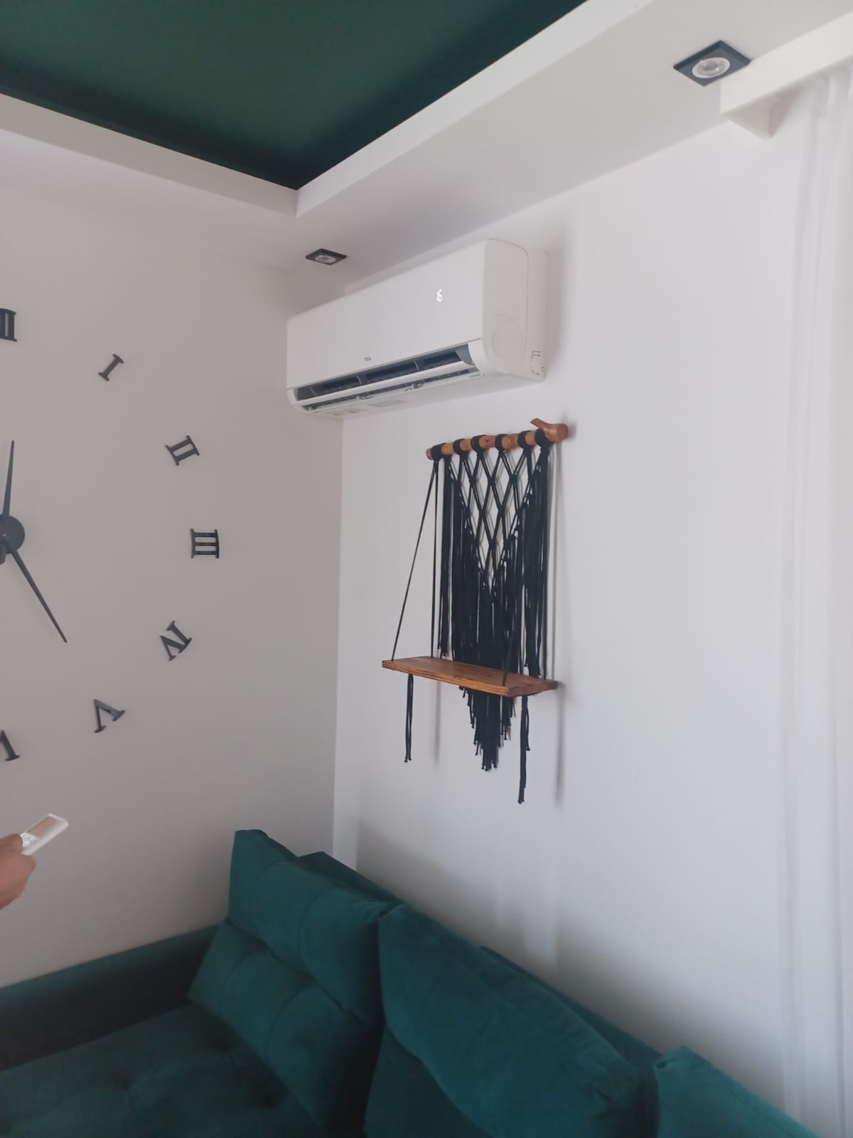 Nowoczesny biały klimatyzator na ścianie w pokoju z zieloną sofą i dekoracyjną półką. Widoczny pilot w dłoni i duży zegar na ścianie.