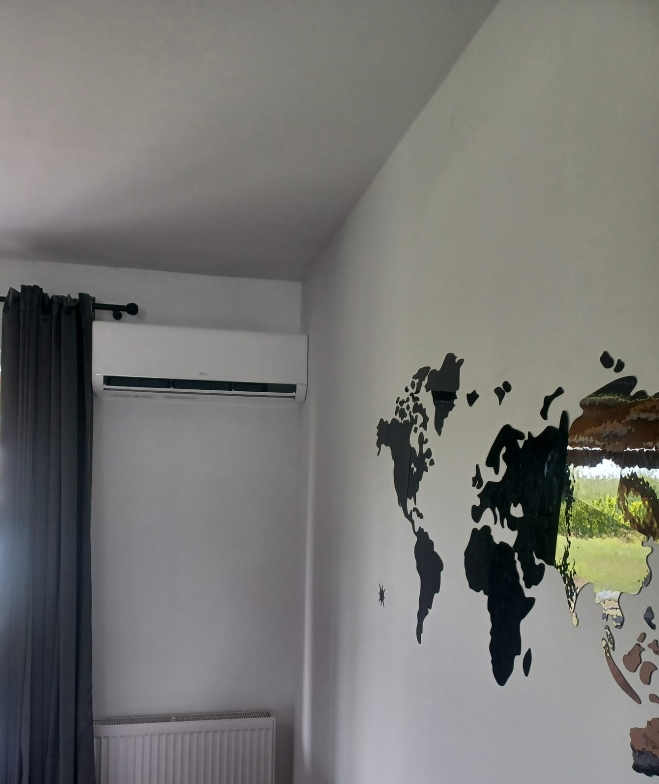 Biała klimatyzacja TCL zamontowana w pokoju z szarymi zasłonami i dekoracyjną mapą świata na ścianie. Widoczny grzejnik pod oknem.