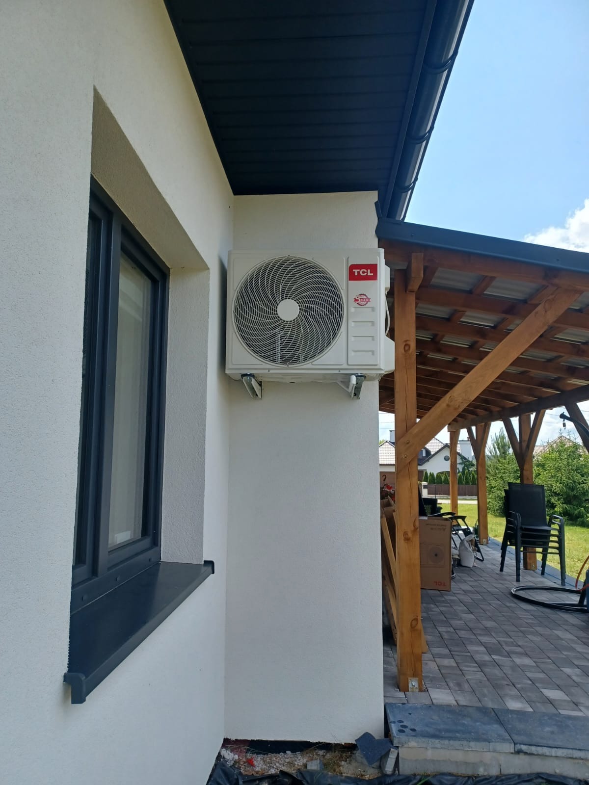 Biały klimatyzator TCL zamontowany na elewacji domu z oknem i drewnianą altaną w tle, widok z boku, słoneczny dzień.