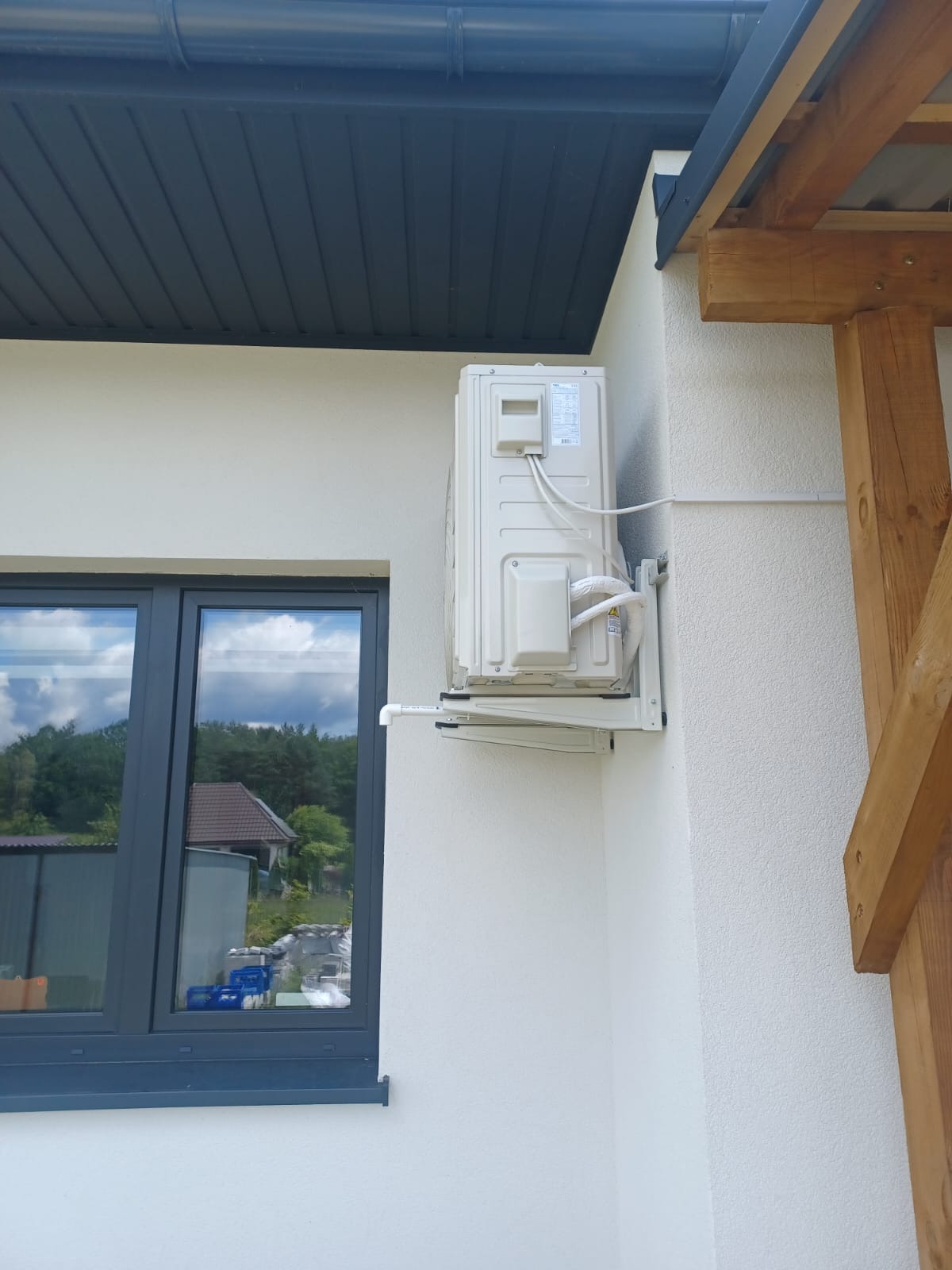 Kompaktowy, kremowy klimatyzator zewnętrzny zamontowany na elewacji budynku obok okna z ciemną ramą. Widoczne rury i kable prowadzone po ścianie. Dach z orynnowaniem.