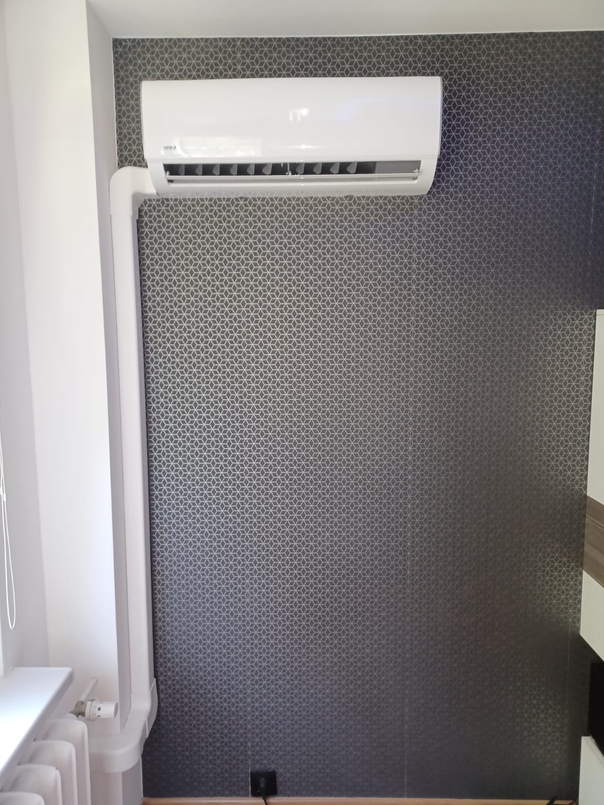 Biała klimatyzacja VIVAX na ścianie z geometrycznym wzorem. Widoczne białe rury odprowadzające. Instalacja w rogu pomieszczenia.