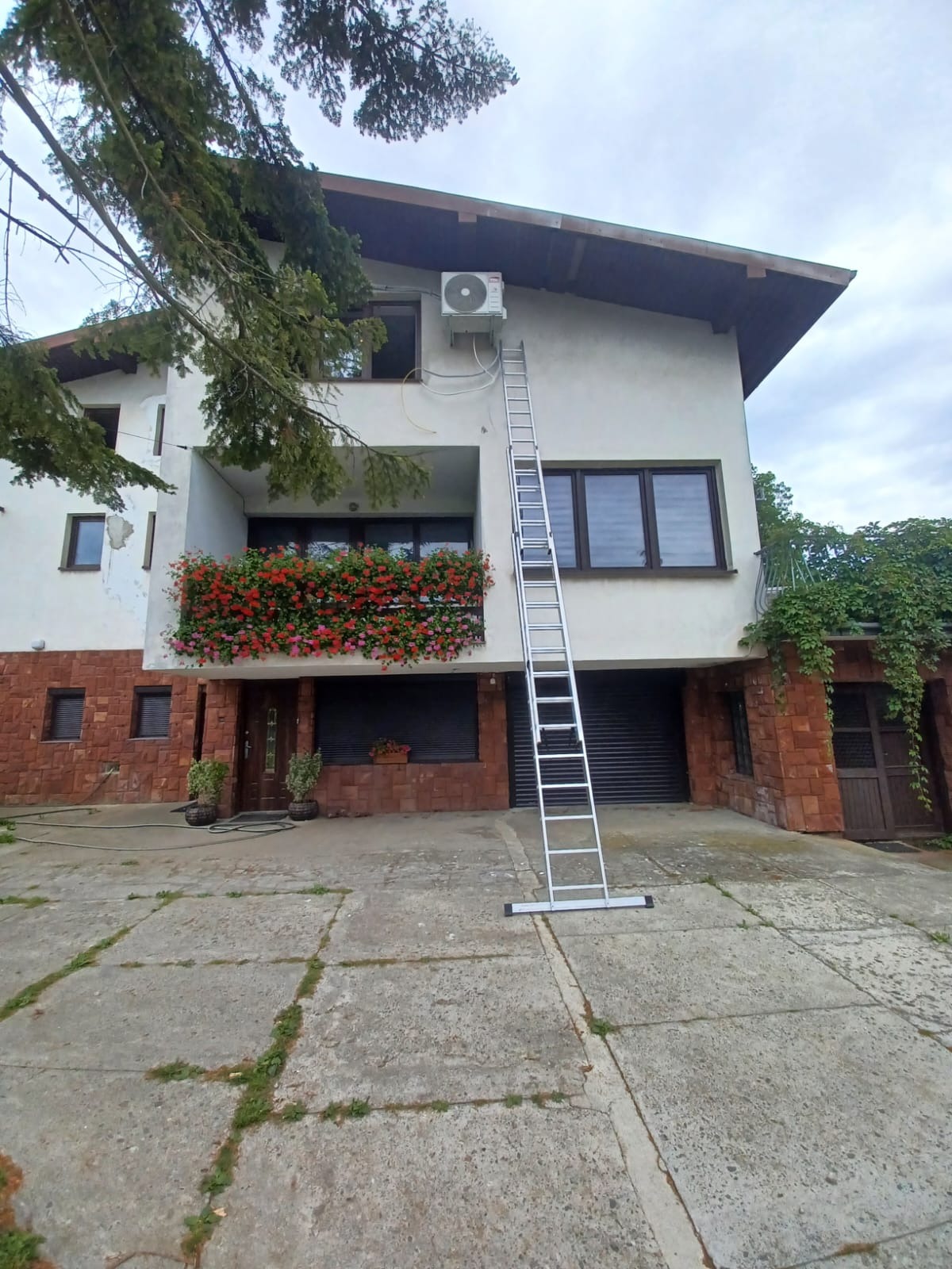 Biały dom z czerwonej cegły, balkon z kwiatami, drabina oparta o ścianę pod klimatyzatorem, widok z dołu, pochmurne niebo.