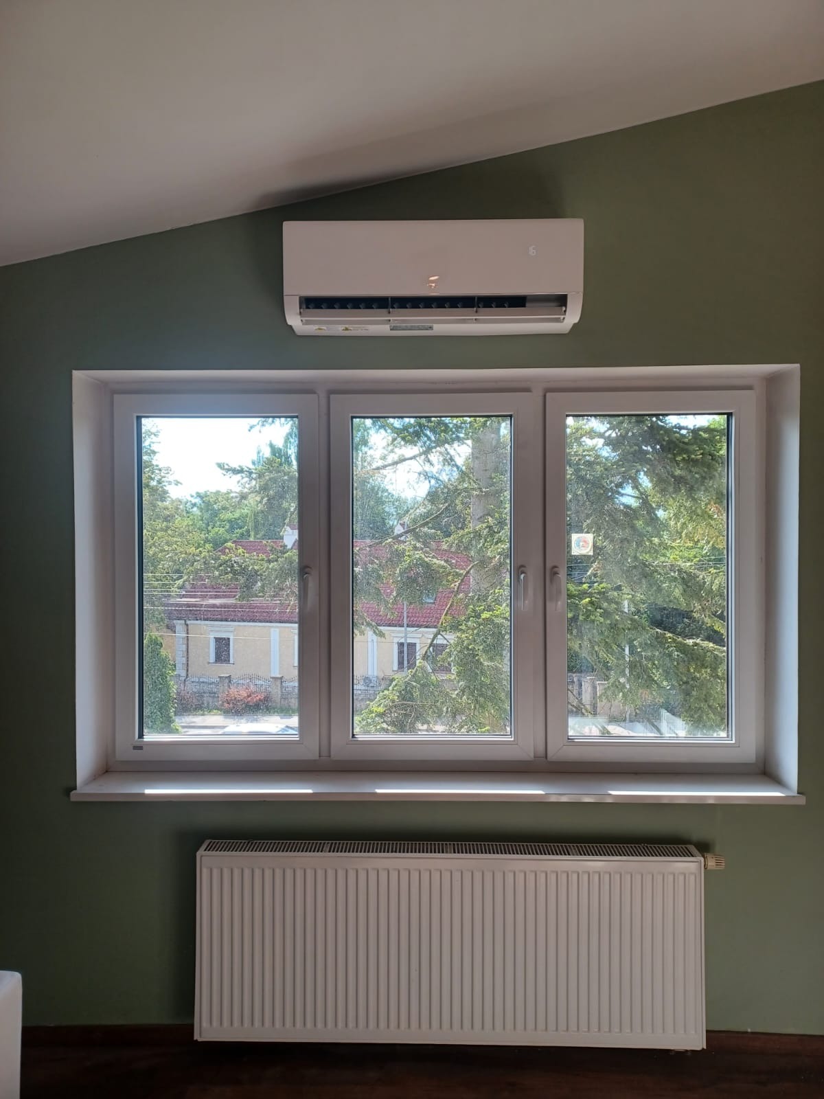 Biały klimatyzator ścienny nad oknem z widokiem na zieleń, pod nim grzejnik. Ściana w odcieniach zieleni. Ujęcie z dołu, widoczny fragment sufitu.
