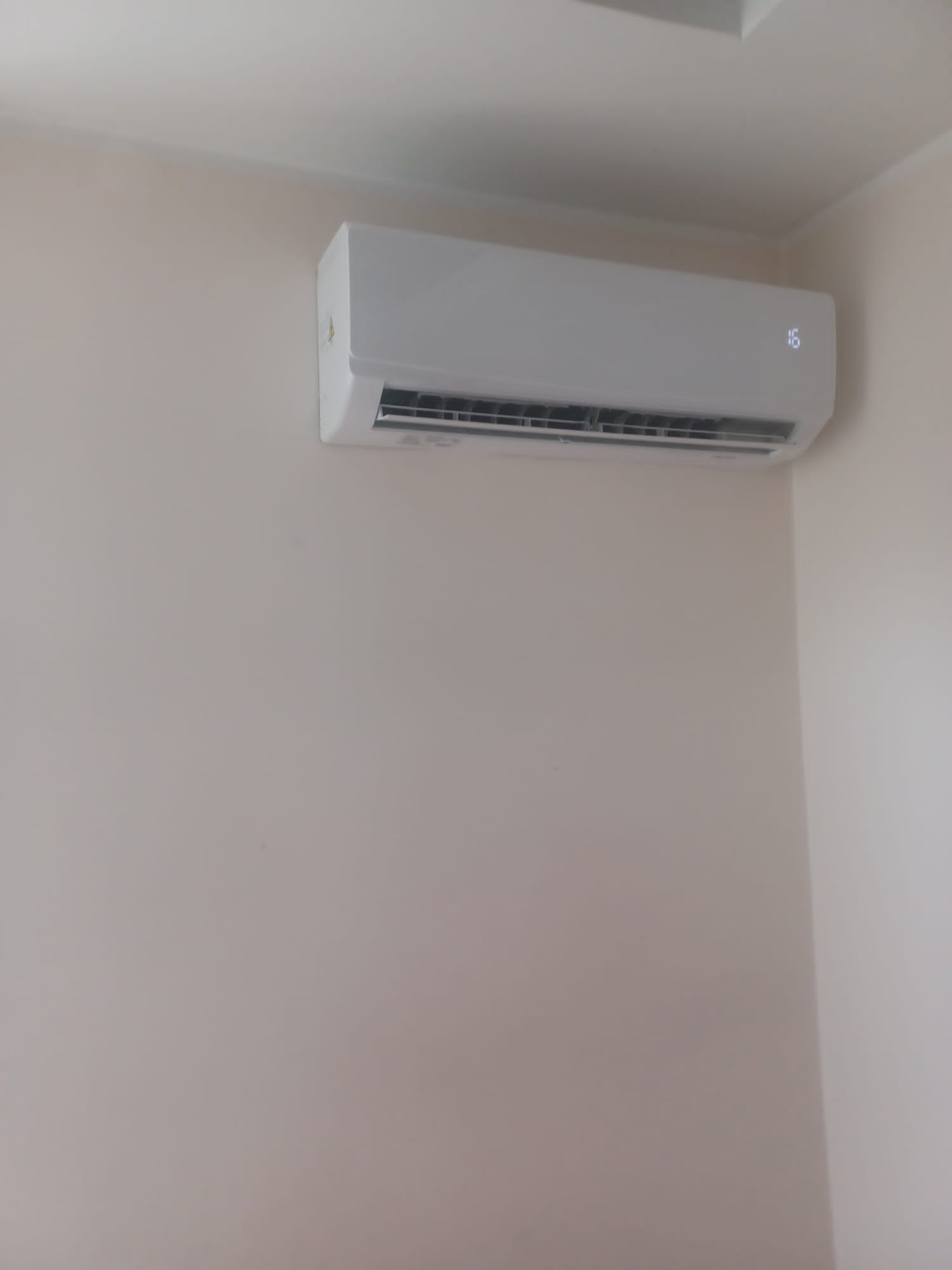 Biały klimatyzator ścienny z wyświetlaczem temperatury 16°C, zamontowany na jasnej ścianie w rogu pomieszczenia. Widok ukośny, oświetlenie naturalne.