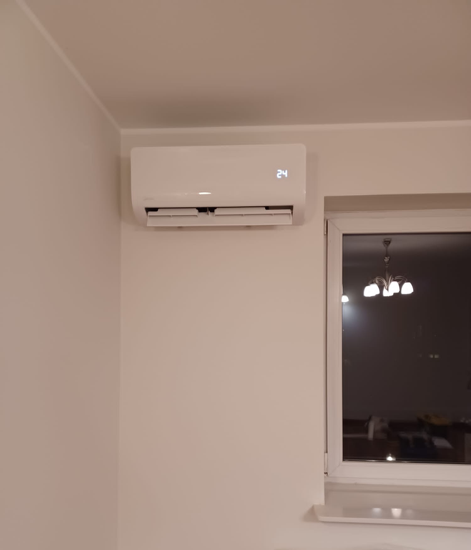Biały klimatyzator ścienny z wyświetlaczem temperatury 24, zamontowany w jasnym pokoju obok okna z widocznym żyrandolem. Minimalistyczny design wnętrza.