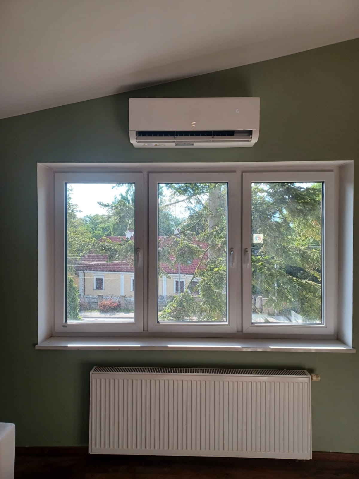 Biała klimatyzacja ścienna nad oknem z widokiem na zieleń, pod oknem biały grzejnik. Ściany w odcieniu zieleni, ukośny sufit.