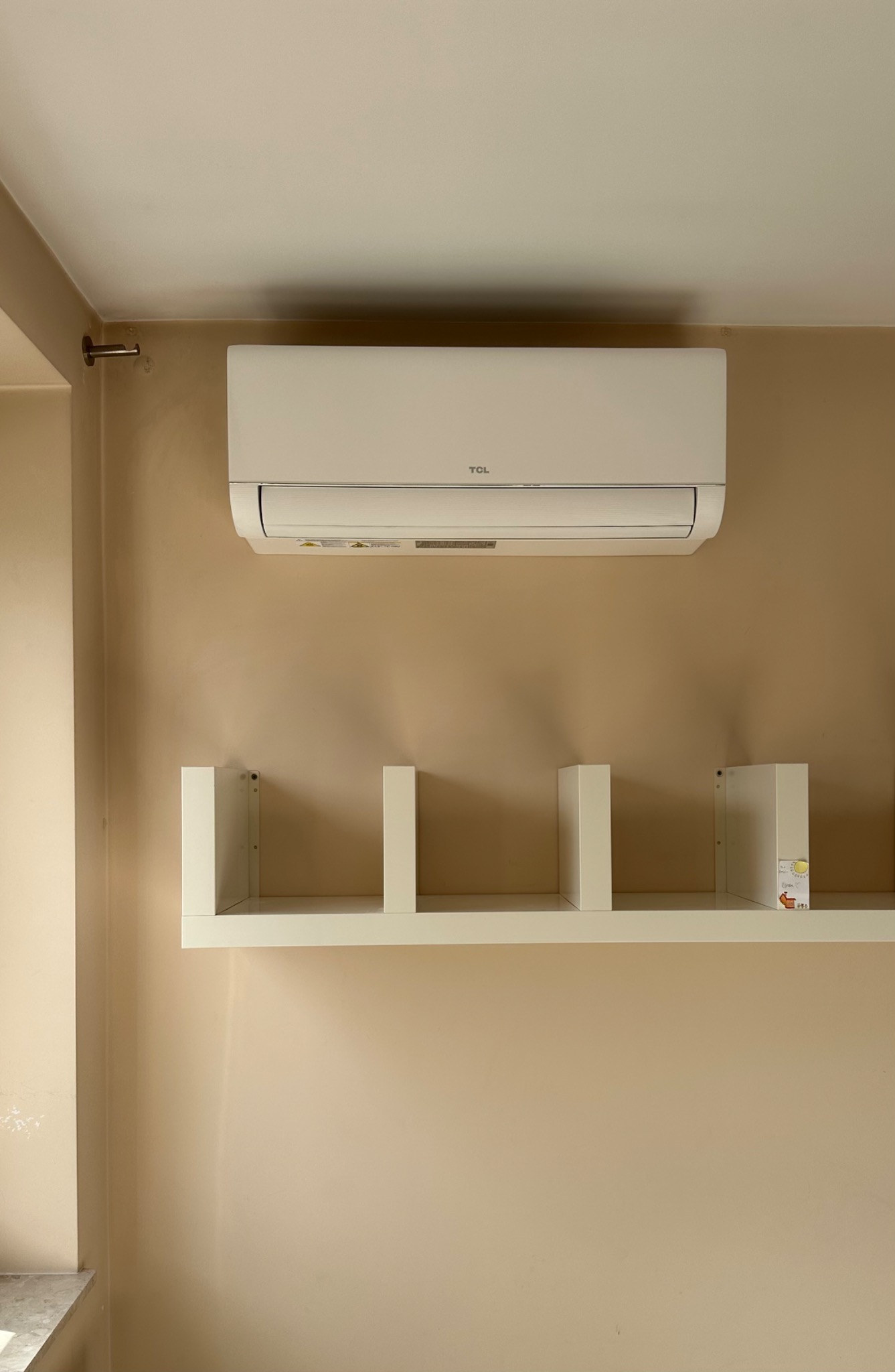 Biały klimatyzator TCL zamontowany na ścianie w beżowym odcieniu, nad białą półką z trzema przegródkami. Widok z przodu, neutralne oświetlenie.