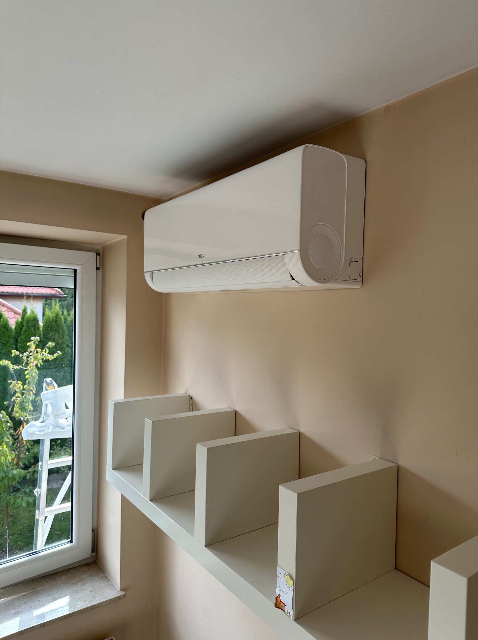 Biały klimatyzator TCL zamontowany na beżowej ścianie w pokoju, widoczne białe półki i okno z widokiem na zieleń. W tle drabina.