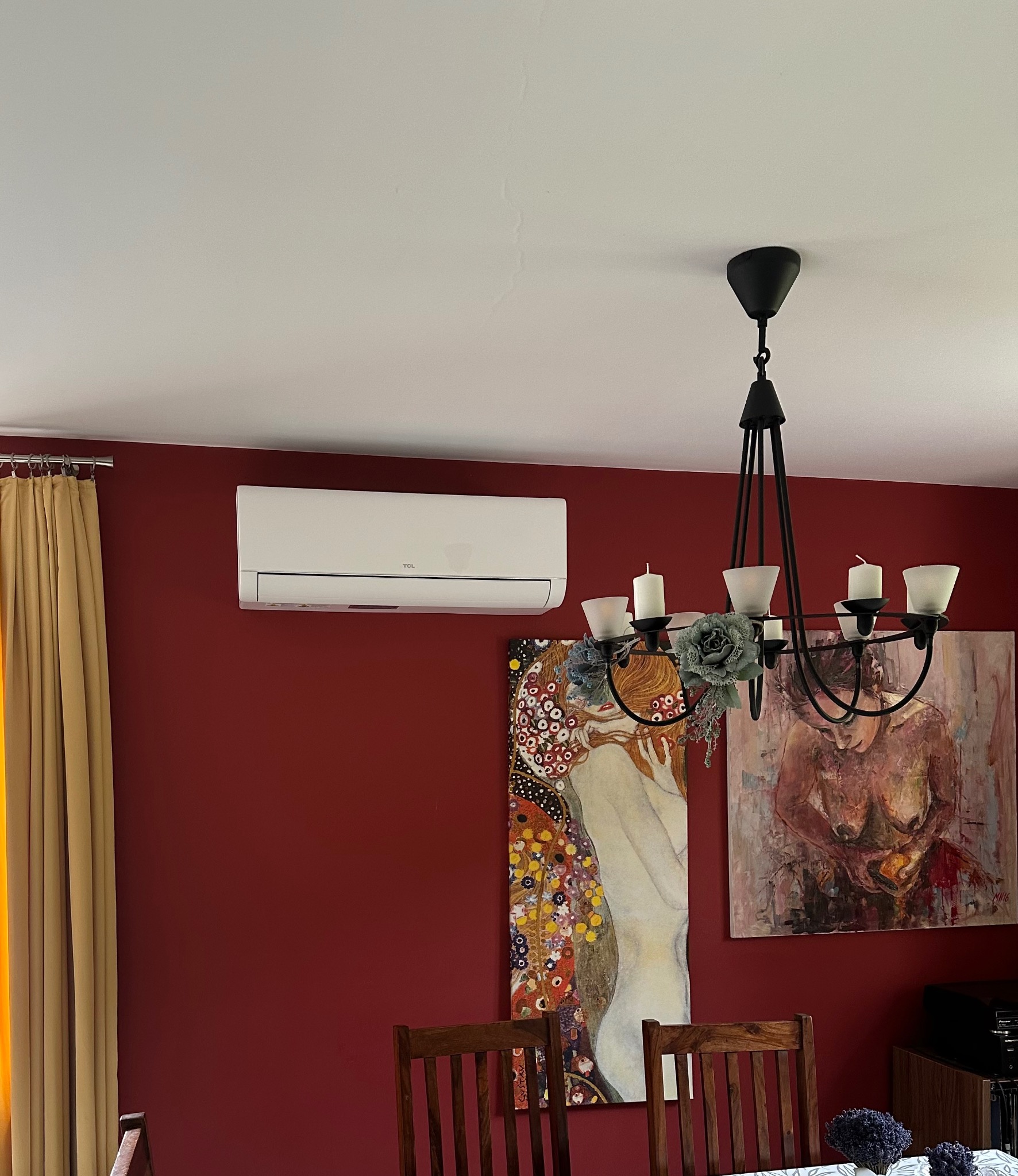 Biała jednostka klimatyzacji TCL na bordowej ścianie w salonie, pod sufitem, z widocznym żyrandolem i obrazami. Widok na wnętrze z drewnianymi krzesłami.