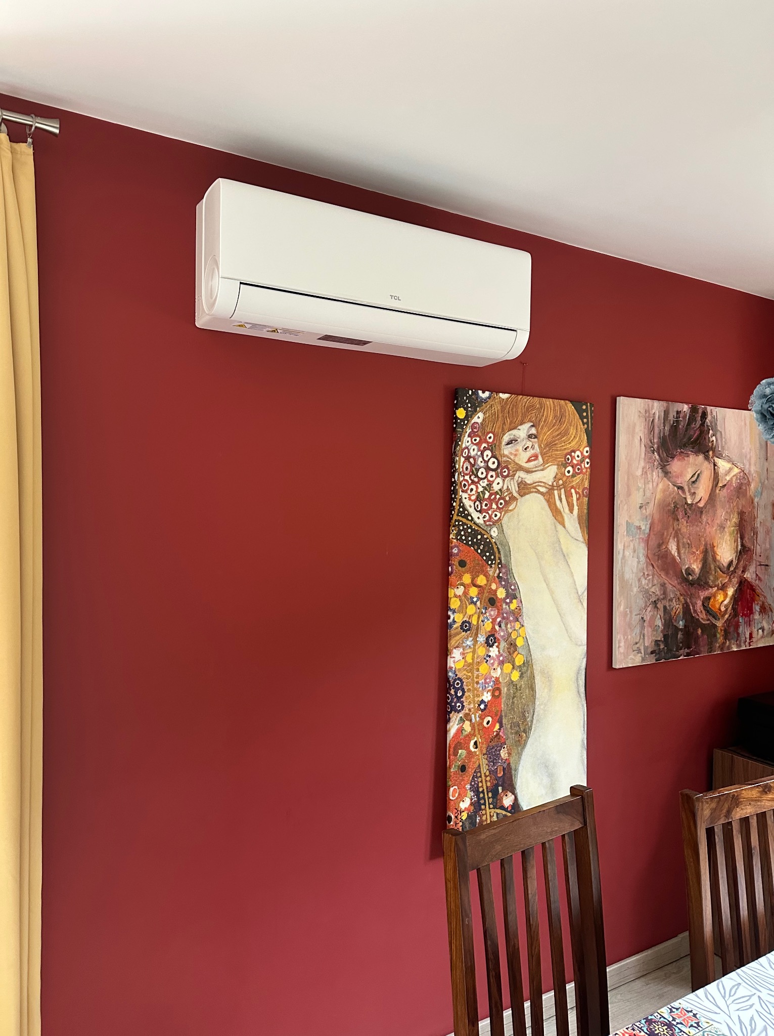 Biały klimatyzator TCL na bordowej ścianie, pod nim obrazy w stylu Klimta. Widoczne krzesła i fragment stołu. Domowe wnętrze z klimatyzacją.