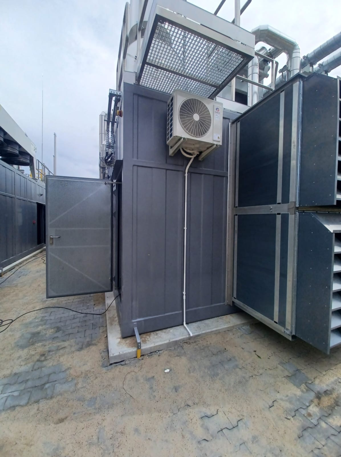 Zewnętrzna instalacja klimatyzacyjna z jednostką na szarej obudowie, widoczne rury i elementy konstrukcyjne. Otwarta metalowa obudowa z kratką wentylacyjną.