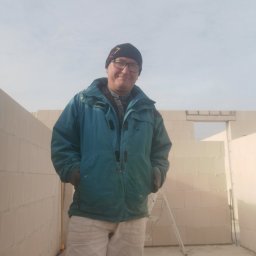 Michał Magulski - Mężczyzna w roboczym ubraniu, na tle surowych ścian z bloczków, w niedokończonym pomieszczeniu z drabiną. W tle błękitne niebo.