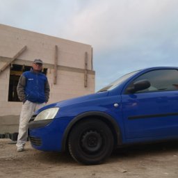 Michał Magulski - Mężczyzna w ubraniu roboczym stoi obok niebieskiego auta na tle ściany budynku w trakcie budowy, widoczne drewniane elementy konstrukcyjne okien.