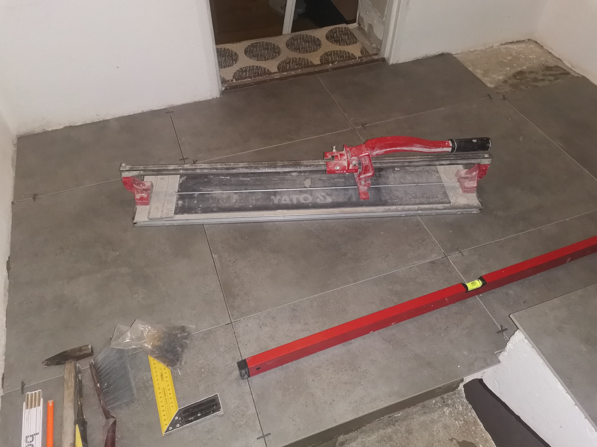 Układanie szarych płytek podłogowych z użyciem poziomicy, przecinarki YATO i narzędzi glazurniczych. Widoczne krzyżyki dystansowe.