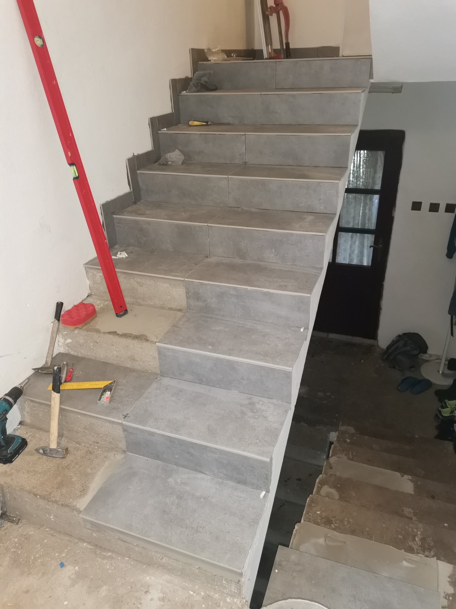 Kładzenie szarych płytek na schodach. Widoczny poziomica, młotek, wiertarka i inne narzędzia. Częściowo wykończone schody prowadzą w górę.