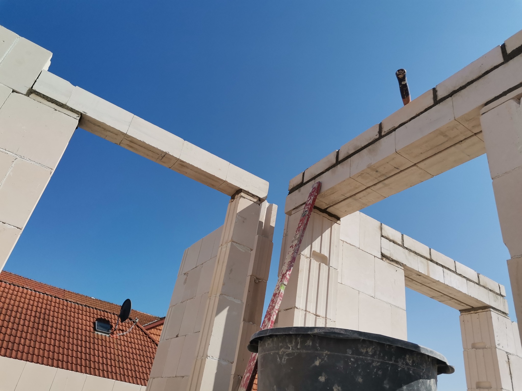 Wznoszenie ściany z bloczków z betonu komórkowego, widok z dołu na fragment konstrukcji, na tle błękitnego nieba. Widoczna poziomica i wiadro.