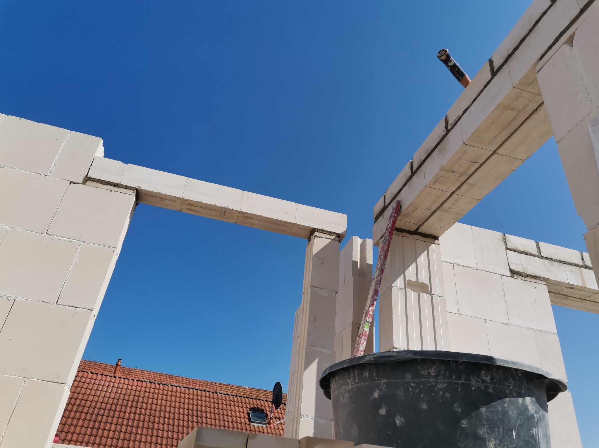 Wznoszenie ścian z białych bloczków silikatowych, widok z dołu na konstrukcję z poziomnicą, kubeł budowlany i fragment dachu w tle, błękitne niebo.