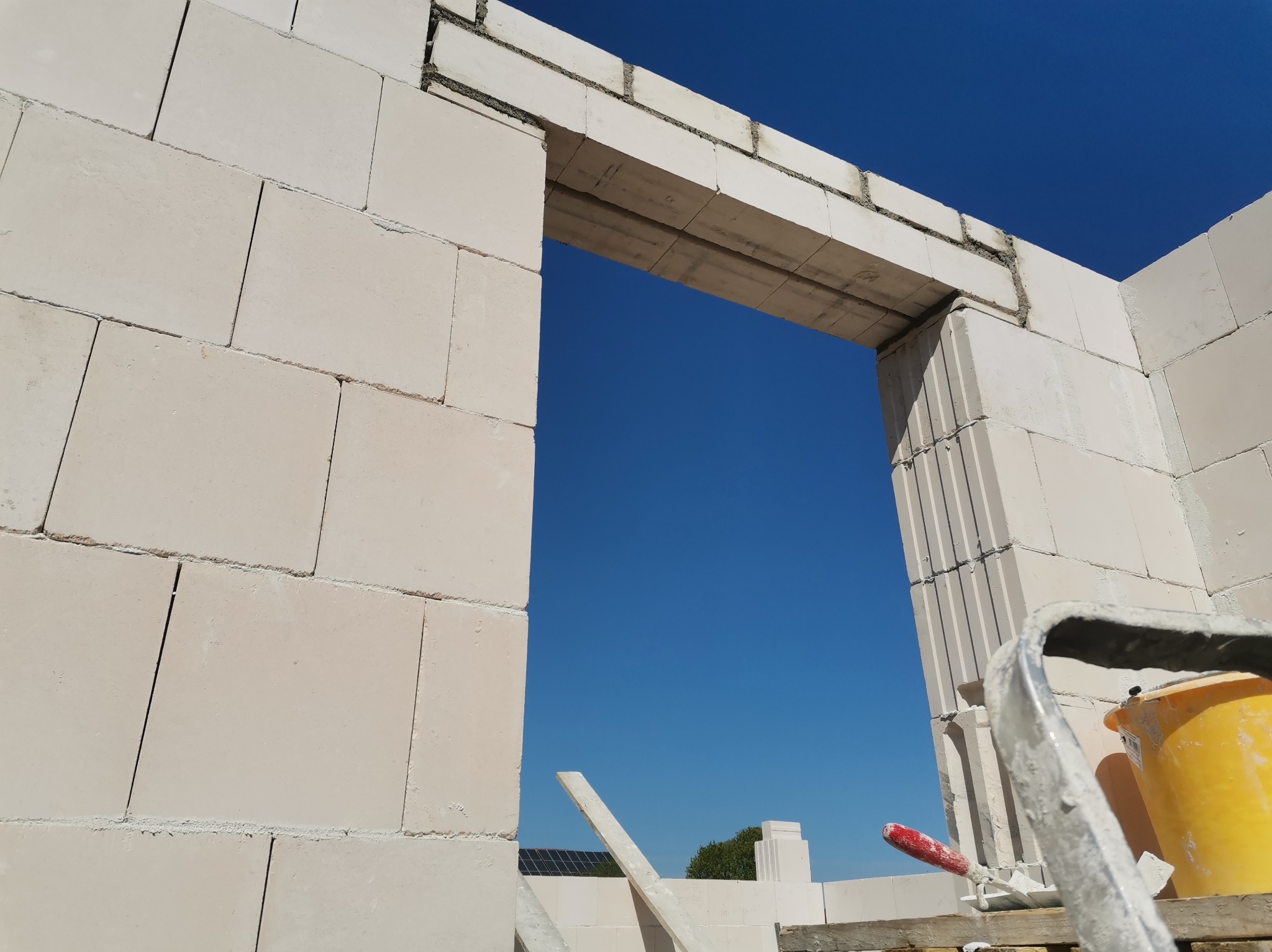 Wznoszenie ściany z bloczków betonu komórkowego, widoczny fragment otworu drzwiowego. Na pierwszym planie wiadro i kielnia. Błękitne niebo w tle.