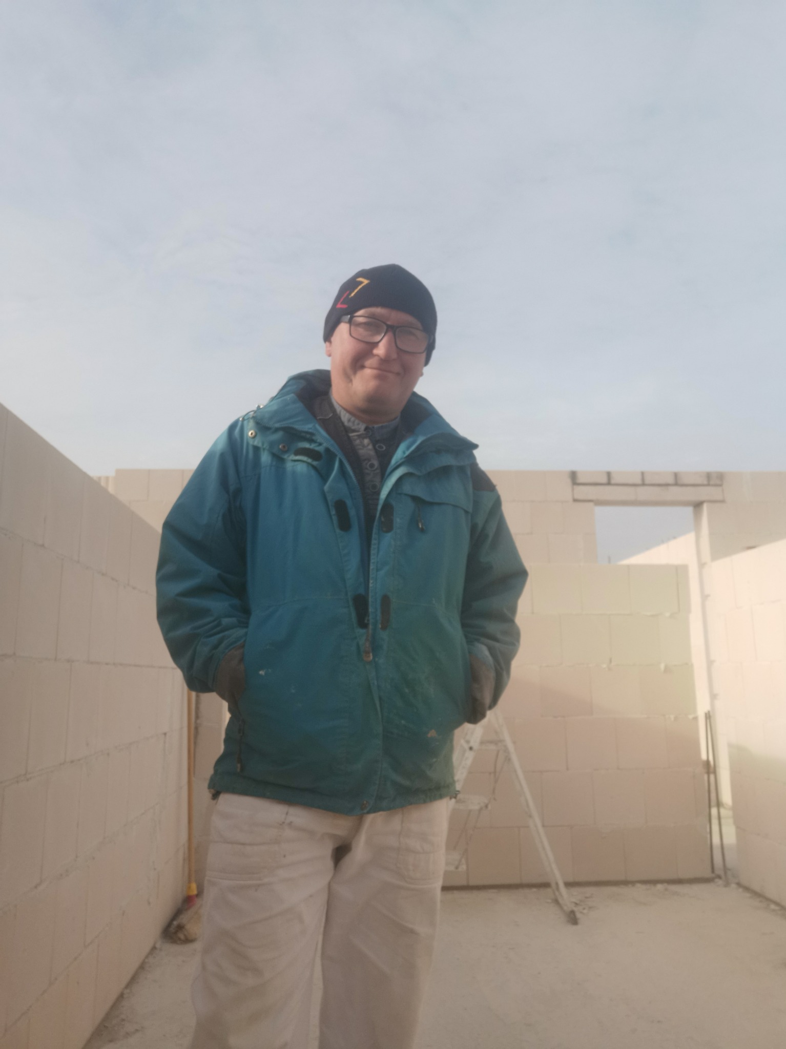 Mężczyzna w roboczym ubraniu, na tle surowych ścian z bloczków, w niedokończonym pomieszczeniu z drabiną. W tle błękitne niebo.