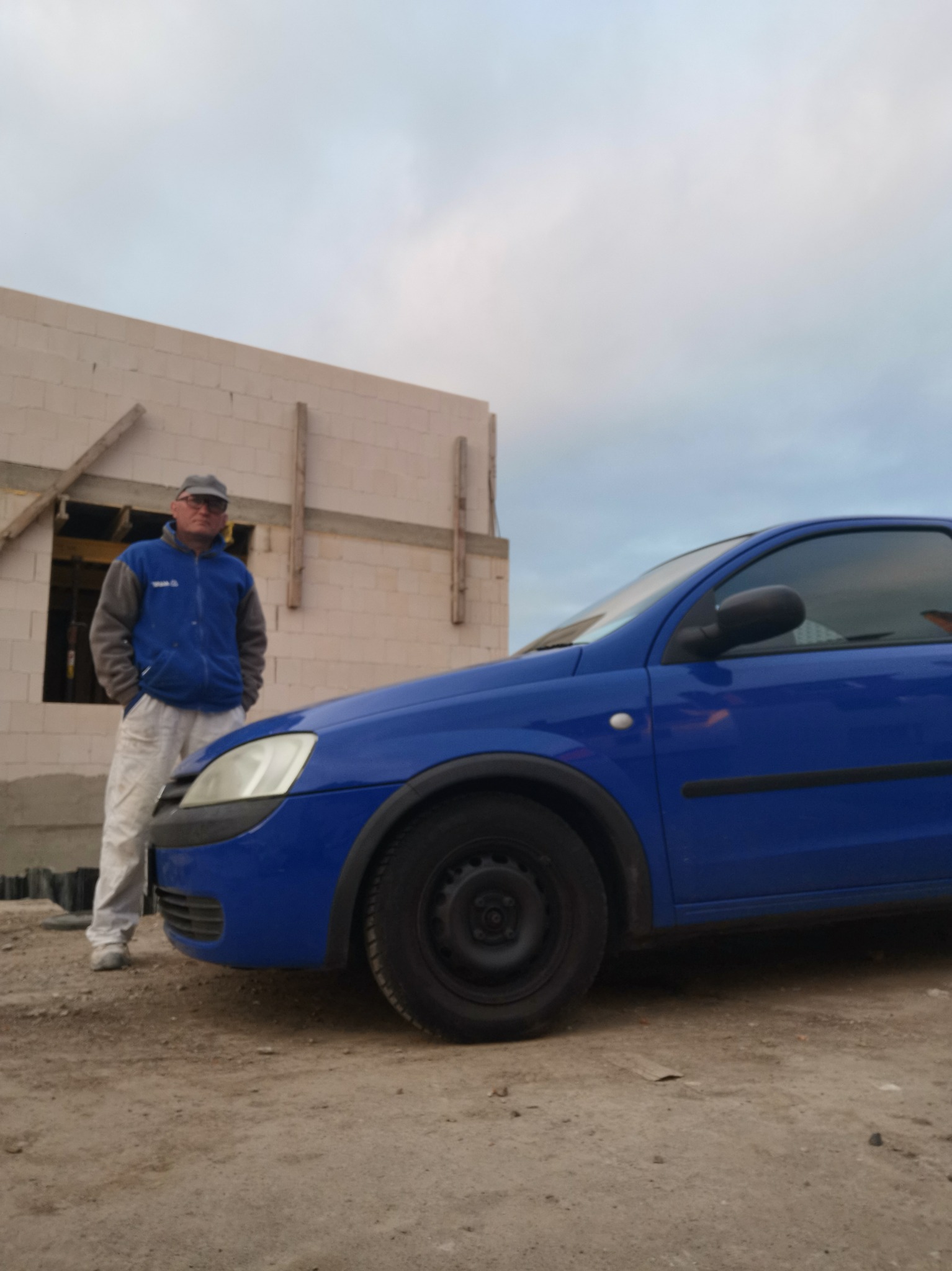 Mężczyzna w ubraniu roboczym stoi obok niebieskiego auta na tle ściany budynku w trakcie budowy, widoczne drewniane elementy konstrukcyjne okien.