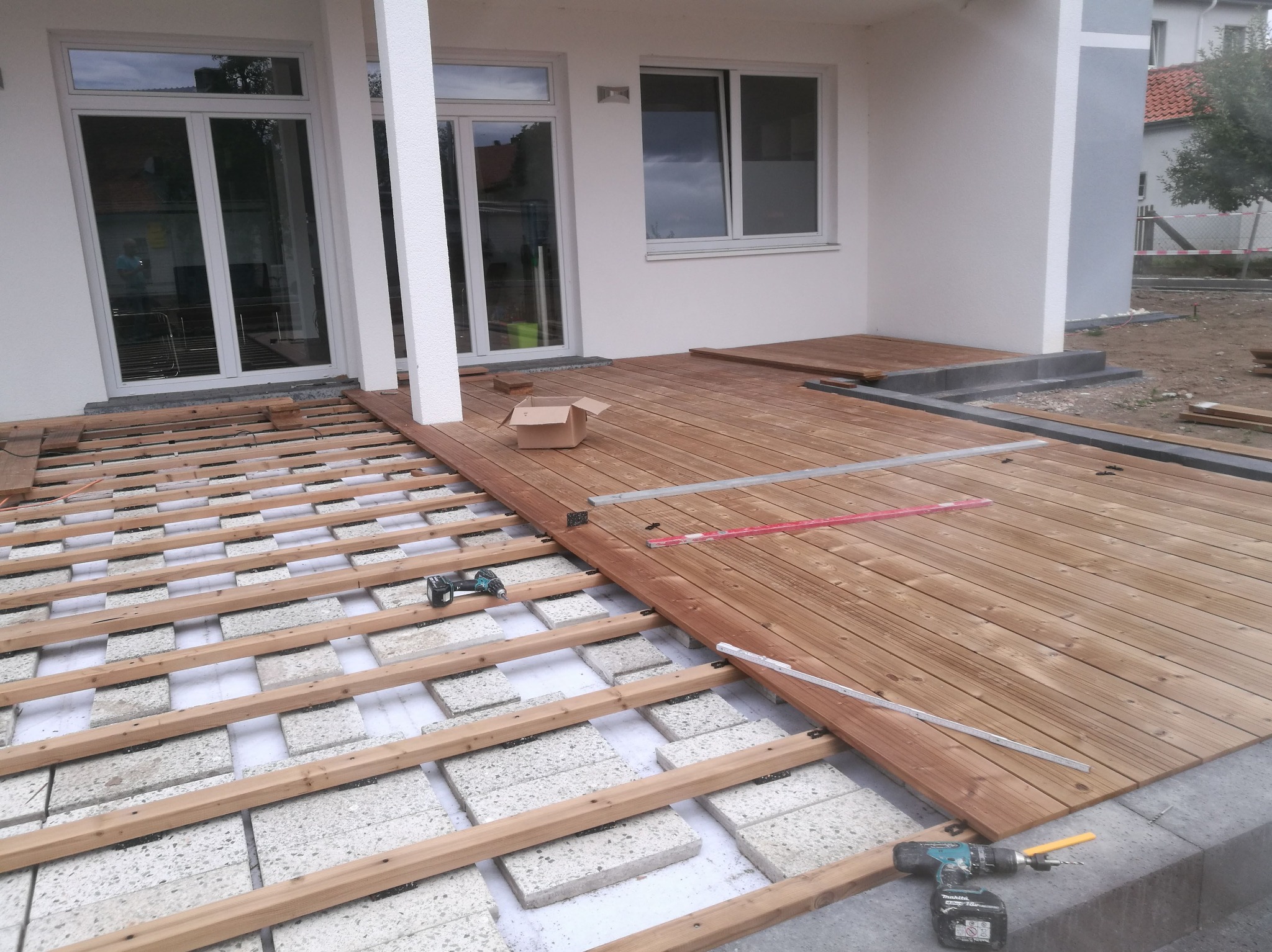 Montaż drewnianego tarasu przy białym budynku. Widoczne narzędzia, deski i podkonstrukcja. Częściowo ułożona podłoga tarasowa z widocznym systemem mocowania.