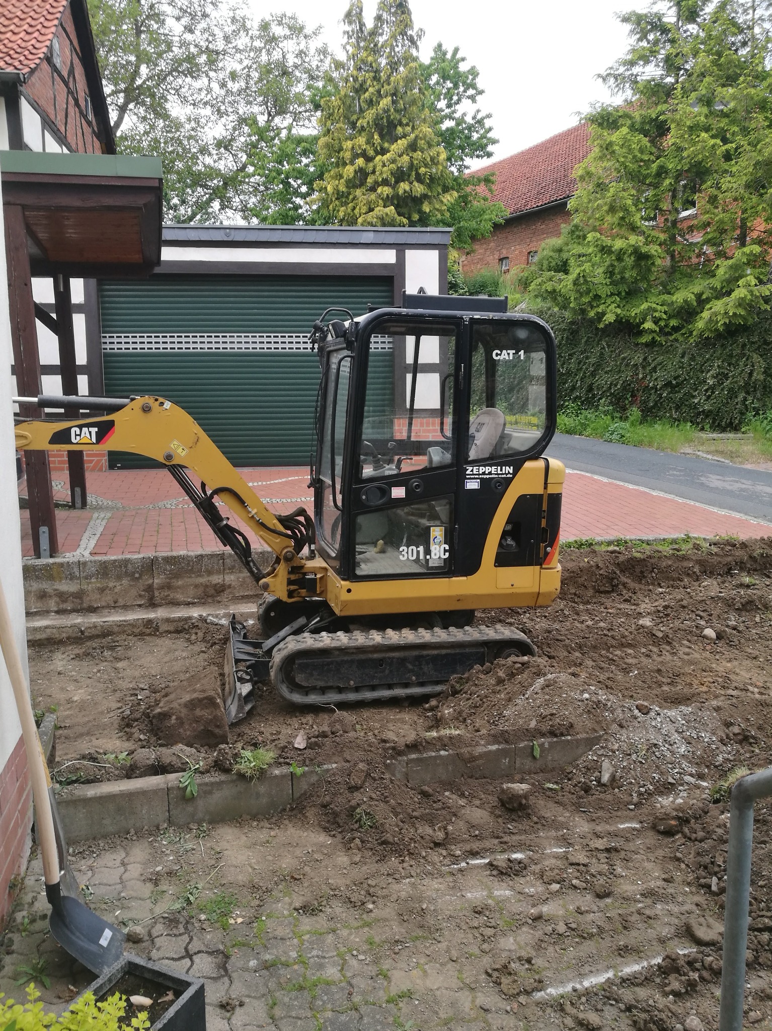 Żółto-czarna mini koparka CAT 301.8C na placu budowy przed domem. Widoczna ziemia i wykopane fundamenty. W tle garaż z zielonymi drzwiami.