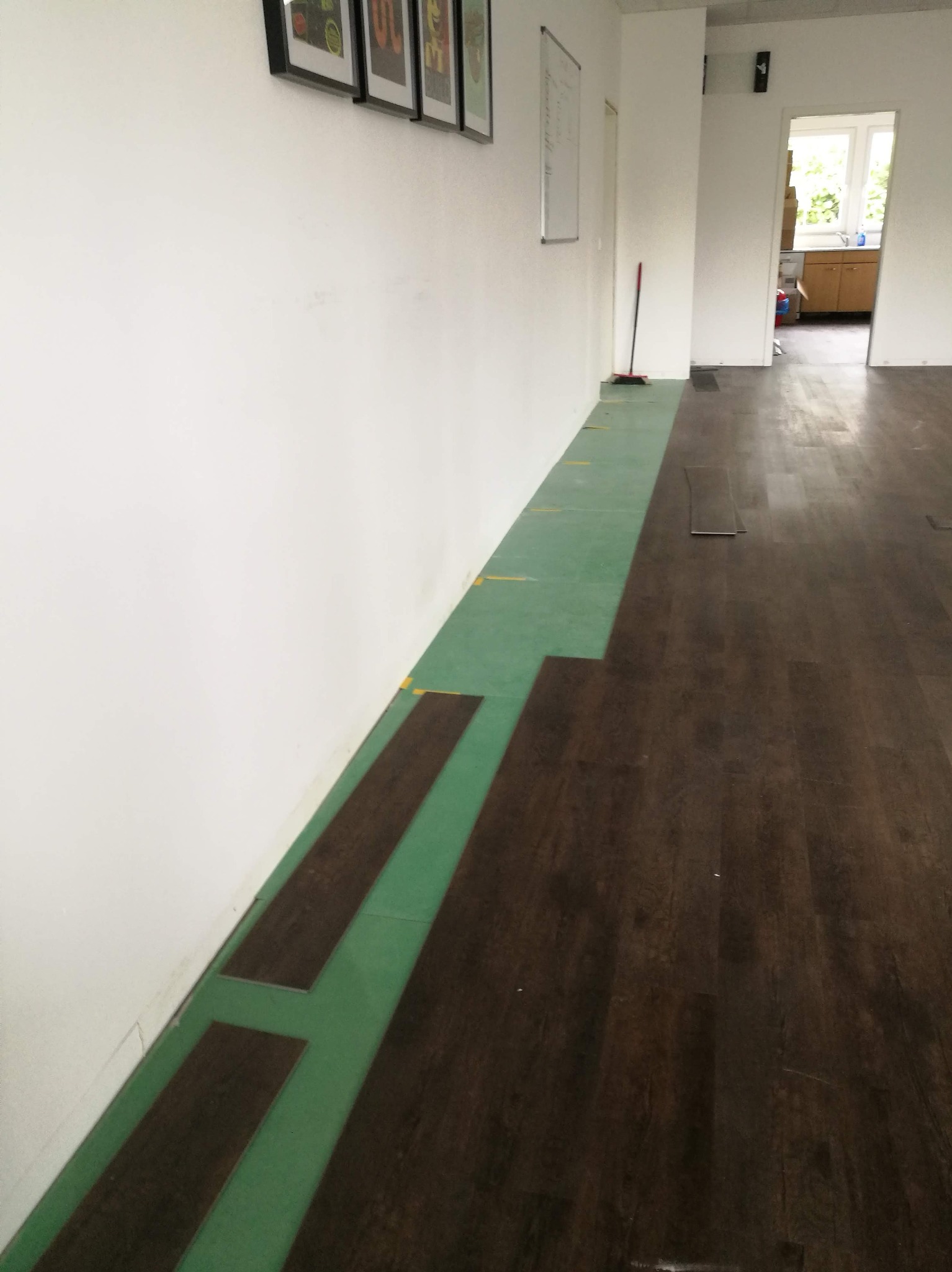 Układanie paneli podłogowych w długim pokoju, widoczna zielona podkładka i ciemne panele, częściowo ułożone. W tle białe ściany i otwarte drzwi do kuchni.