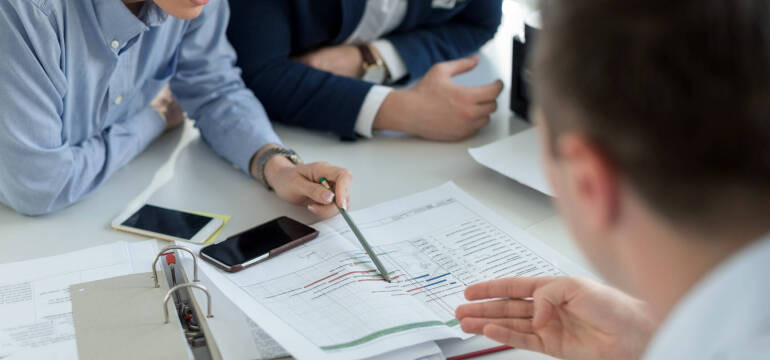 Spotkanie biznesowe: analiza wykresów z użyciem ołówka, telefony na stole, widoczne fragmenty ubrań uczestników, jasne oświetlenie, dokumenty w segregatorze.